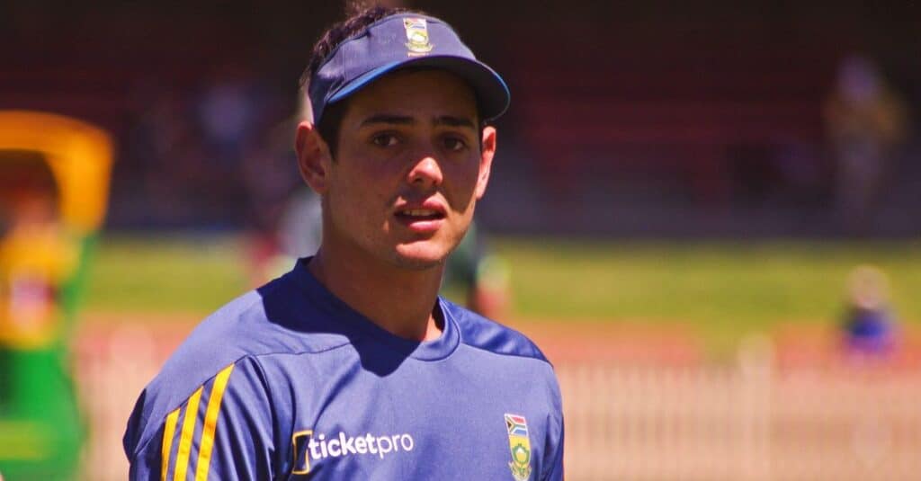 Quinton de Kock wearing a blue and yellow shirt and cap, smiling at the camera.